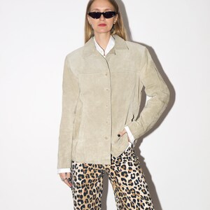 Pu&ograve; includere: Giacca in pelle scamosciata beige chiaro con chiusura a bottoni e colletto. La giacca &egrave; indossata sopra una camicia bianca con colletto e abbinata a pantaloni con stampa leopardata. La modella indossa occhiali da sole neri.