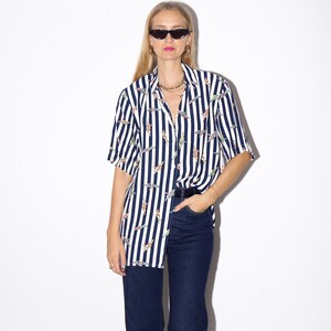 May include: A short-sleeved, button-down shirt with vertical navy and white stripes. The shirt features a pattern of colorful, abstract figures. The shirt is paired with dark blue jeans. The model is wearing sunglasses and gold hoop earrings.