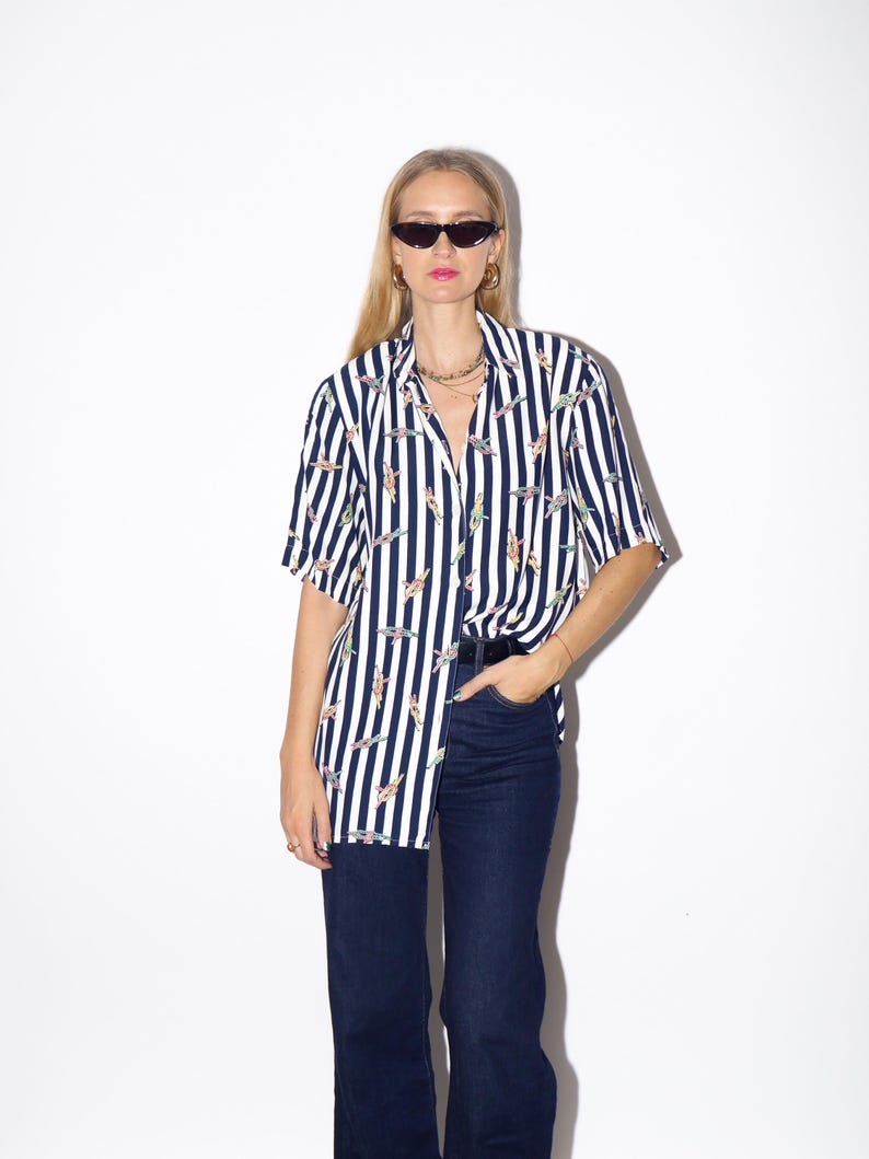 May include: A short-sleeved, button-down shirt with vertical navy and white stripes. The shirt features a pattern of colorful, abstract figures. The shirt is paired with dark blue jeans. The model is wearing sunglasses and gold hoop earrings.