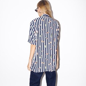May include: A short-sleeved shirt with vertical navy blue and white stripes. The shirt features a pattern of colorful, abstract shapes. The person in the image is wearing dark blue pants and sunglasses.