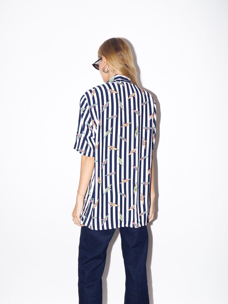 May include: A short-sleeved shirt with vertical navy blue and white stripes. The shirt features a pattern of colorful, abstract shapes. The person in the image is wearing dark blue pants and sunglasses.