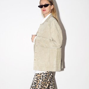Pu&ograve; includere: Giacca in camoscio beige chiaro con bottoni. La giacca &egrave; indossata sopra una camicia bianca con colletto e abbinata a pantaloni con stampa leopardata. La persona indossa occhiali da sole neri a occhi di gatto.