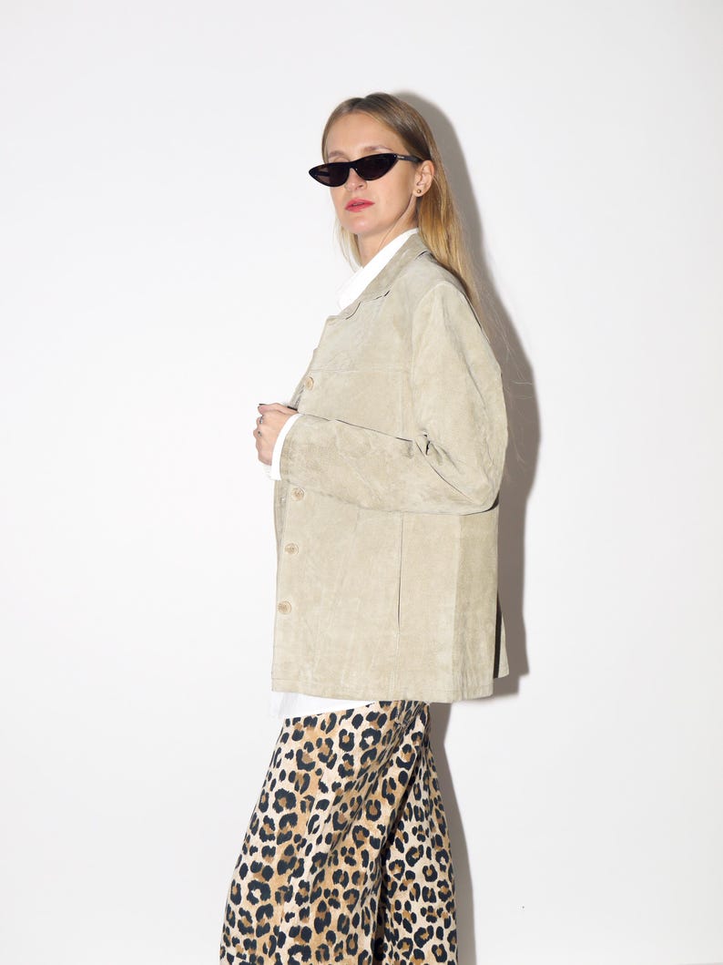 Pu&ograve; includere: Giacca in camoscio beige chiaro con bottoni. La giacca &egrave; indossata sopra una camicia bianca con colletto e abbinata a pantaloni con stampa leopardata. La persona indossa occhiali da sole neri a occhi di gatto.