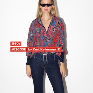 Op de afbeelding: Een vrouw draagt een vintage viscose shirt met paisley print, V-hals en donkerblauwe jeans. Het shirt heeft rode, blauwe en zwarte patronen. De jeans heeft een hoge taille en een zilveren gespriem. Er wordt een zonnebril gedragen.