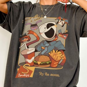 May include: A charcoal grey oversized t-shirt featuring a graphic print of a cartoon character in a uniform, a burger, chips, and a drink. The text reads "Serve with a smile!", "Spudsy's", and "Try the sauce." The shirt has short sleeves and a crew neck.
