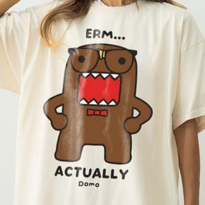 May include: An ivory short-sleeve t-shirt featuring a brown cartoon character wearing black glasses and a red bow tie. The character has a wide open mouth with sharp teeth. Above the character is the text ERM... and below is ACTUALLY Domo.