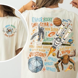 May include: An ivory t-shirt featuring a space-themed graphic. The back displays a rocket, a crab-like creature, a geometric sphere, and text including "Grace Rocky Save Star You Sleep I Watch". The front chest shows a smaller matching graphic.