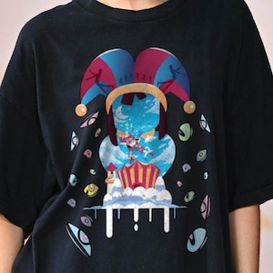 May include: A black t-shirt featuring a graphic print of a jester hat in blue and magenta, positioned above a surreal scene with a circus tent, clouds, and a crescent moon. The design is surrounded by multiple floating eyes in various colours.