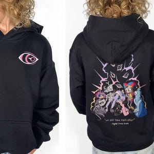 May include: A black hooded sweatshirt featuring a small pink and white eye graphic on the front chest. The back displays a colorful illustration of multiple cartoon characters with the text "we still have each other" and "Digital Circus Finale".