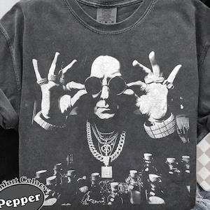 Peut inclure: Un t-shirt à manches courtes gris anthracite avec une impression graphique en noir et blanc représentant une personne portant des lunettes rondes et plusieurs colliers superposés, les mains levées près du visage. Finition délavée.