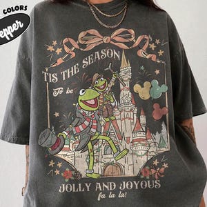 May include: A charcoal gray short-sleeve t-shirt featuring a graphic of Kermit the Frog and Fozzie Bear in front of a castle. The text reads 'TIS THE SEASON To be JOLLY AND JOYOUS fa la la!' with a ribbon, stars, and Mickey-shaped balloons.