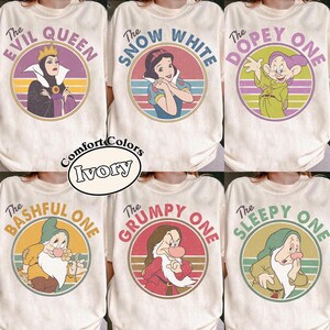 Peut inclure: Six t-shirts ivoire avec des graphismes circulaires rétro de personnages de Blanche-Neige. Chaque t-shirt présente un personnage avec du texte incluant The Evil Queen, The Snow White, The Dopey One, The Bashful One, The Grumpy One et The Sleepy One.