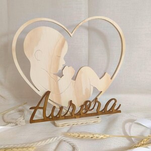 Pode incluir: Decoração de madeira com tema de bebé. Apresenta uma moldura em forma de coração com uma silhueta de bebé no interior. O nome "Aurora" está escrito em itálico abaixo do coração. A decoração é feita de madeira clara e está sobre uma superfície branca.