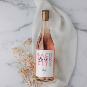May include: A bottle of pink wine with a white label that reads "Bachlorette Barbie" in pink lettering. The bottle is on a white cloth with a sprig of dried flowers.