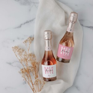 May include: Two small bottles of pink champagne with labels that say "Bachlorette" in pink letters. The bottles are on a white cloth with a sprig of dried flowers.