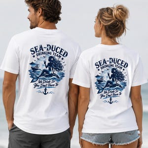 Puede incluir: Una camiseta blanca con un gráfico azul en la espalda. El diseño incluye una sirena sentada sobre una roca, un barco y el texto "SEA-DUCED DRINKING TEAM" y "We Drink So You Don't Have To" con un icono de ancla en la parte inferior.