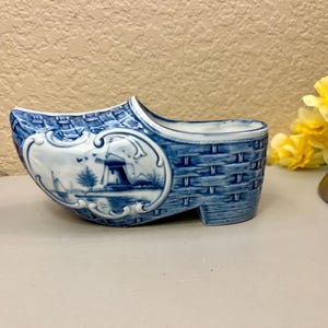 May include: A blue and white ceramic Dutch shoe planter with a windmill scene. The shoe is decorated with a woven pattern and has a small opening at the top for planting.