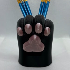 May include: A black paw-shaped desk organizer holding several paint brushes. The organizer has a pink paw pad and toe beans. The paint brushes have blue handles and gold bristles.
