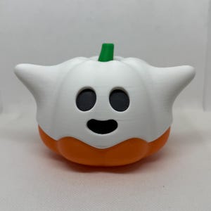 A Halloween decoration combining a white ghost and an orange pumpkin. The ghost has black eyes and a smiling mouth. A green stem is on top. The item is 3D-printed. The decoration is suitable for Halloween.