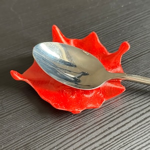 Könnte beinhalten: Ein Edelstahl-Löffel liegt auf einer leuchtend roten Löffelablage. Die Löffelablage hat eine einzigartige, organische Form mit erhöhten Kanten. Der Löffel ist mittig auf der Ablage platziert und reflektiert das Licht. Die Löffelablage besteht aus einem festen Material.