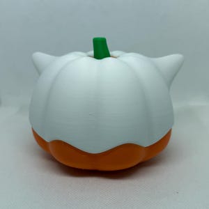 May include: A white and orange pumpkin-shaped ornament with a green stem. The pumpkin has a rounded shape with a white top and orange base. The item has two small, pointed white ears on either side.