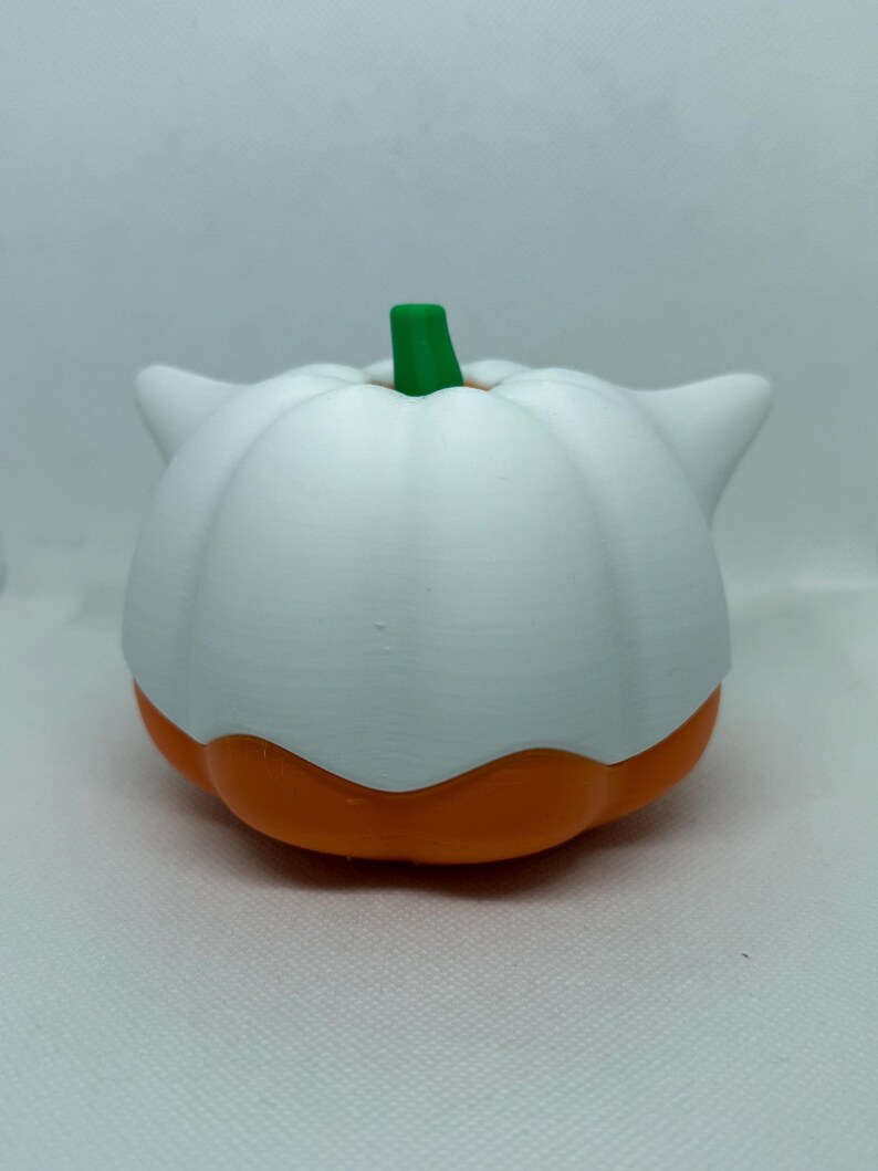 May include: A white and orange pumpkin-shaped ornament with a green stem. The pumpkin has a rounded shape with a white top and orange base. The item has two small, pointed white ears on either side.