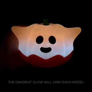 May include: A pumpkin-shaped light with a smiling face. The light has a white and orange gradient, a red base, and a green stem. The text at the bottom reads, "THE GRADIENT GLOW WILL VARY EACH MODEL."