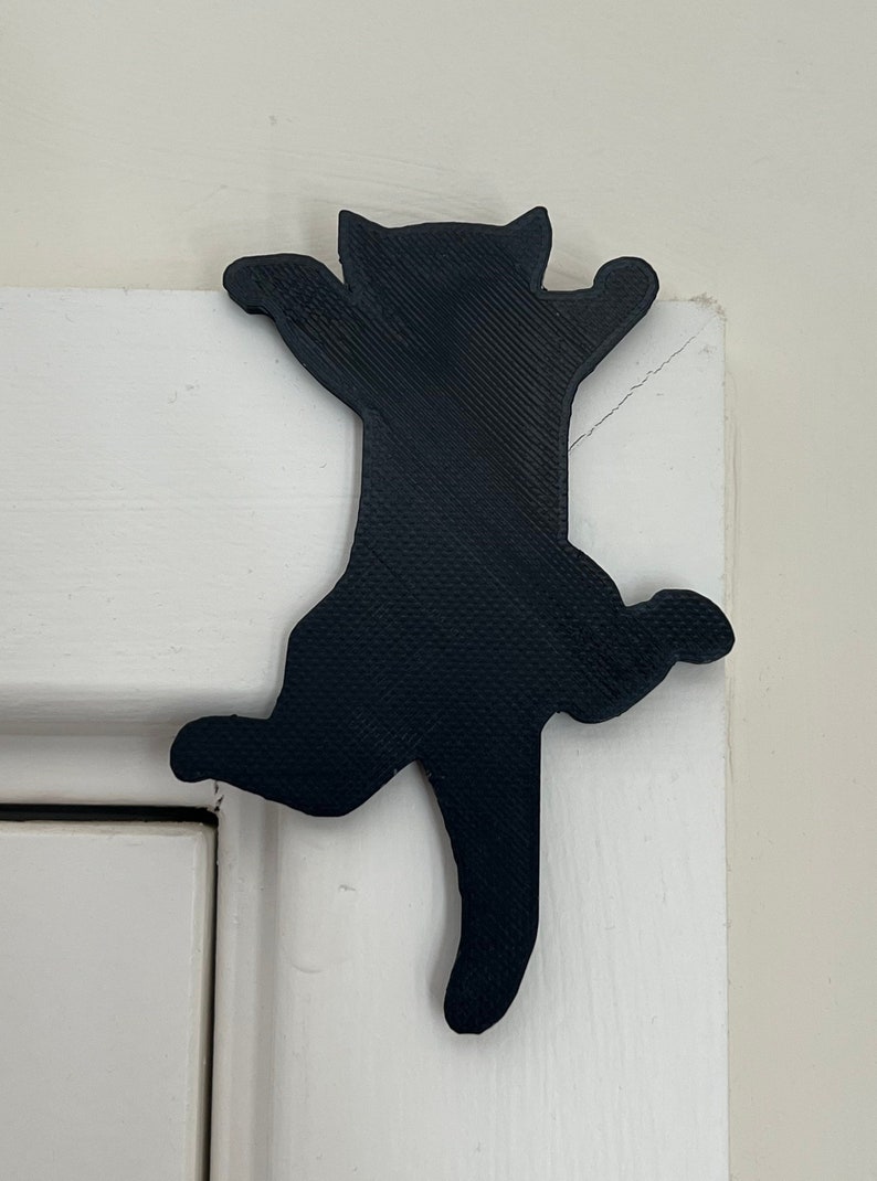 Op de afbeelding: Een zwart 3D-geprint katten silhouet, hangend aan een witte deur. De kat bevindt zich in een klimhouding met uitgestrekte poten.