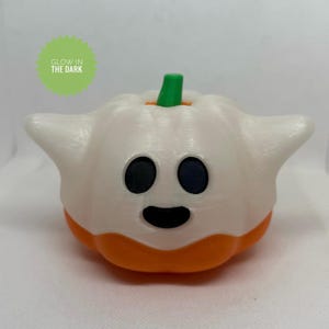 May include: A white and orange Halloween decoration in the shape of a ghost-pumpkin hybrid. The ghost-pumpkin has black eyes and a smiling mouth. The stem is green. The text "GLOW IN THE DARK" is in a green circle.