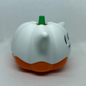May include: A white and orange Halloween pumpkin decoration with a green stem. The pumpkin has a ghost-like design with cut-out eyes and a small horn. The base of the pumpkin is orange.