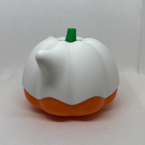 May include: A white and orange pumpkin-shaped container with a green stem. The container has a spout and is likely used for pouring liquids. The design is reminiscent of a gourd, with a ribbed texture on the white upper portion.