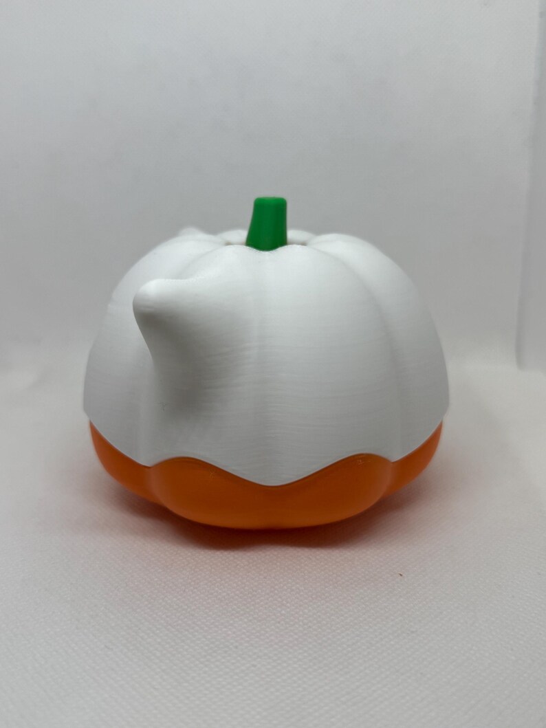 May include: A white and orange pumpkin-shaped container with a green stem. The container has a spout and is likely used for pouring liquids. The design is reminiscent of a gourd, with a ribbed texture on the white upper portion.
