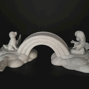 May include: A light-colored, speckled figurine featuring a central arched bridge connecting two cloud-shaped bases. On one side, a small dog sits facing the bridge, while on the other side, a small figure with outstretched arms faces the bridge.