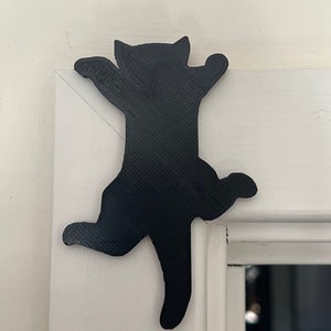 Op de afbeelding: Zwarte silhouet van een kat die een wit deurkozijn beklimt. De kat staat in een speelse houding met zijn poten uitgestrekt.