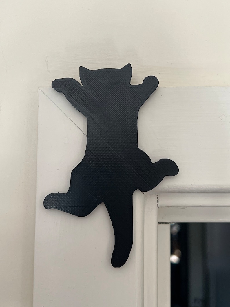 Op de afbeelding: Zwarte silhouet van een kat die een wit deurkozijn beklimt. De kat staat in een speelse houding met zijn poten uitgestrekt.