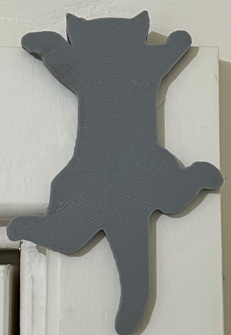 Op de afbeelding: Een grijze 3D-geprinte katten-silhouet deurstop. De kat staat in een springende pose met zijn poten omhoog.