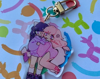 Op de afbeelding: Transparante acryl sleutelhanger met een anime-stijl illustratie van twee figuren die elkaar omhelzen. Eén figuur heeft paars haar, de ander roze. De sleutelhanger heeft een goudkleurige sluiting. Achtergrond met ballonnen.