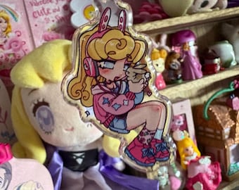 Op de afbeelding: Een doorzichtige acryl sleutelhanger met een cartoonillustratie van een blond personage met roze koptelefoon met konijnenoren, gekleed in een roze jas met blauwe sterren, een blauwe rok en roze sneakers, terwijl het een klein bruin konijntje vasthoudt.