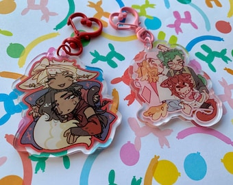 Op de afbeelding: Twee heldere acryl sleutelhangers met cartoon illustraties van personages. Eén sleutelhanger heeft een rode hartvormige sluiting, de andere een roze hartvormige sluiting. De sleutelhangers liggen op een kleurrijke achtergrond.