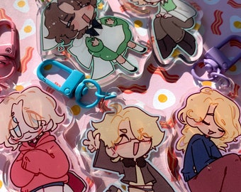 Op de afbeelding: Een verzameling heldere acryl sleutelhangers met cartoon illustraties van personages in verschillende poses. De sleutelhangers hebben gekleurde metalen sluitingen in groen, roze en blauw. De achtergrond is een roze patroon met spek- en spiegelei ontwerpen.
