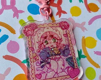 Op de afbeelding: Een roze acryl sleutelhanger met een cartoon illustratie van een meisje met roze haar en een hartvormige sluiting. De illustratie is omlijst met rozen en harten, tegen een kleurrijke achtergrond van ballonfiguren.