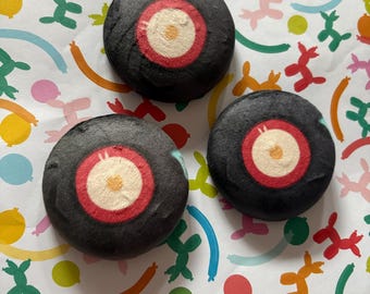 Op de afbeelding: Drie ronde, zwarte bruisballen met een centraal doelwitontwerp met een witte binnenring, een rode ring en een gele stip in het midden. Het oppervlak van elke bruisbal is glad en mat, met een consistente ronde vorm en een duidelijk patroon.