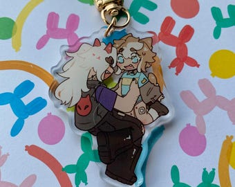 Op de afbeelding: Een heldere acryl sleutelhanger met een cartoon illustratie van twee personages die elkaar omarmen. De sleutelhanger heeft een goudkleurige sluiting. De achtergrond is een wit oppervlak met kleurrijke ballonhond illustraties.