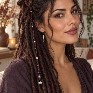Könnte beinhalten: Ein Haaraccessoire bestehend aus einer dunkelroten Kordel, die mit verschiedenen kleinen Perlen in Rosa-, Grau- und Schwarztönen verflochten ist. Das Accessoire endet in einem silberfarbenen Metallanhänger in Form eines Ginkgoblattes für Dreadlocks.