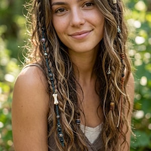 May include: A woman with long, wavy, light brown hair styled with braids and decorative beads. The braids are adorned with bone, wooden, and metal accents. She wears a brown top and smiles at the camera, with a blurred green background.