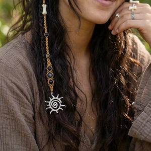 May include: A woman with long, dark wavy hair decorated with a macrame hair wrap. The wrap includes black beads, bone-shaped beads, and a sun-shaped pendant. She wears rings and a brown top.