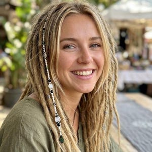 Könnte beinhalten: Eine lächelnde Person mit blonden Dreadlocks, die mit dekorativen Perlen verziert sind. Die Dreadlocks sind mit einer Strähne aus weißen, schwarzen und grünen Perlen versehen. Die Person trägt ein grünes Hemd.