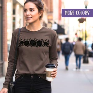 May include: An espresso brown long-sleeve shirt featuring a black floral graphic across the chest. The shirt has a crew neckline and is paired with black pants. The text "ESPRESSO" is visible on the left and "MORE COLORS" is in the top right corner.
