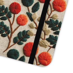 Puede incluir: Primer plano de una cubierta de cuaderno con diseño floral bordado. El diseño presenta flores y hojas naranjas, rojas, verdes y beige sobre un fondo beige. Una banda elástica negra asegura el cuaderno.