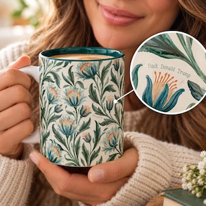 May include: A ceramic mug with a white base and a repeating pattern of teal and green botanical flowers and leaves. The interior rim is dark teal. The text 'Fuck Donald Trump' is printed in small black letters among the floral design.
